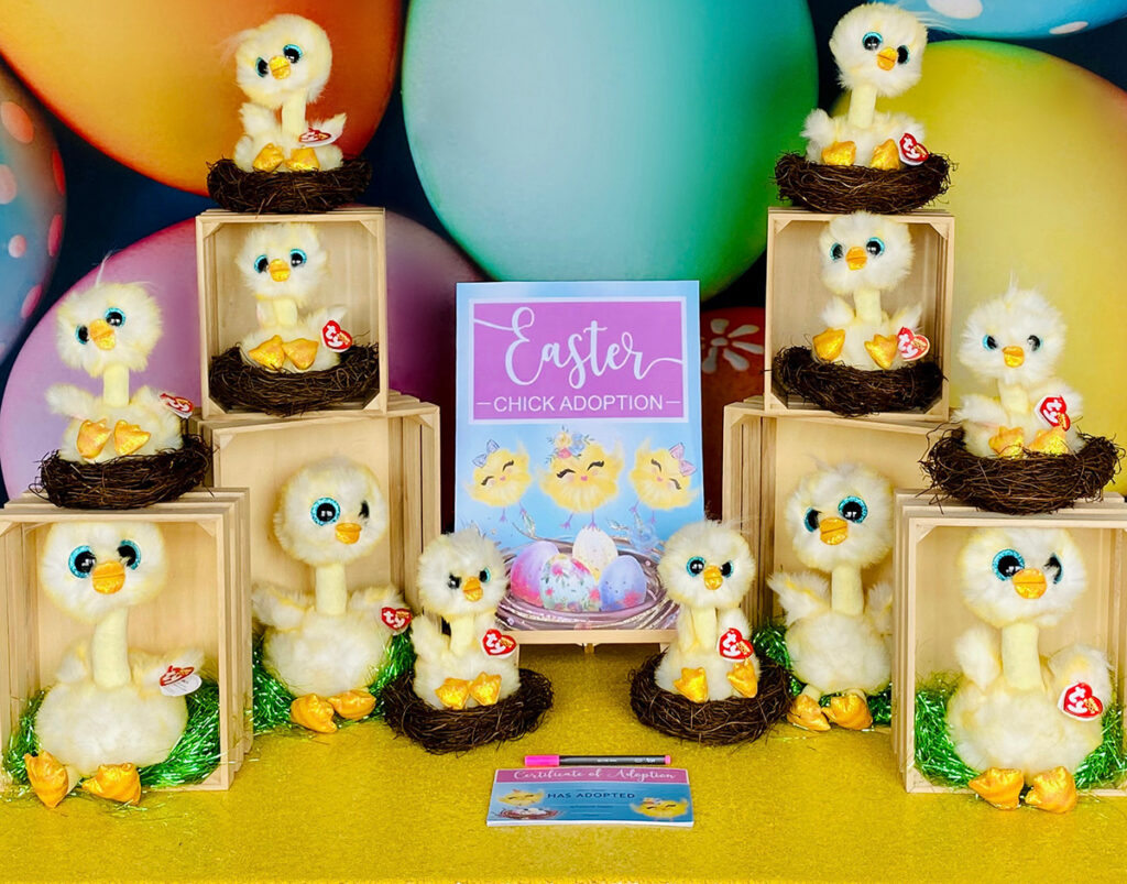 Close-up of the Easter chick adoption party table featuring plush chicks in nests and crates, a festive backdrop, a party sign, and adoption certificates displayed as part of the setup.