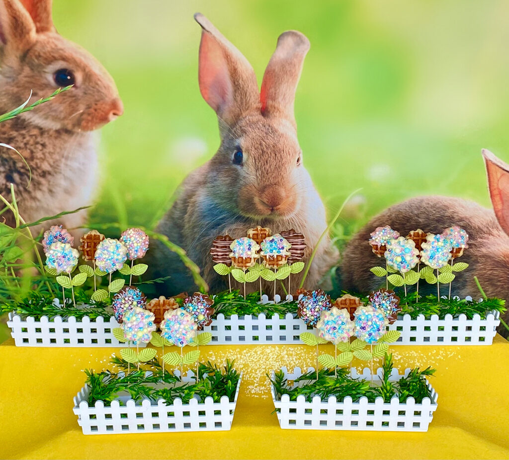 Easter waffle pop party display styled like a spring garden with floral waffle pops in white picket crates, set against a bunny-themed backdrop and yellow sequin tablecloth.
