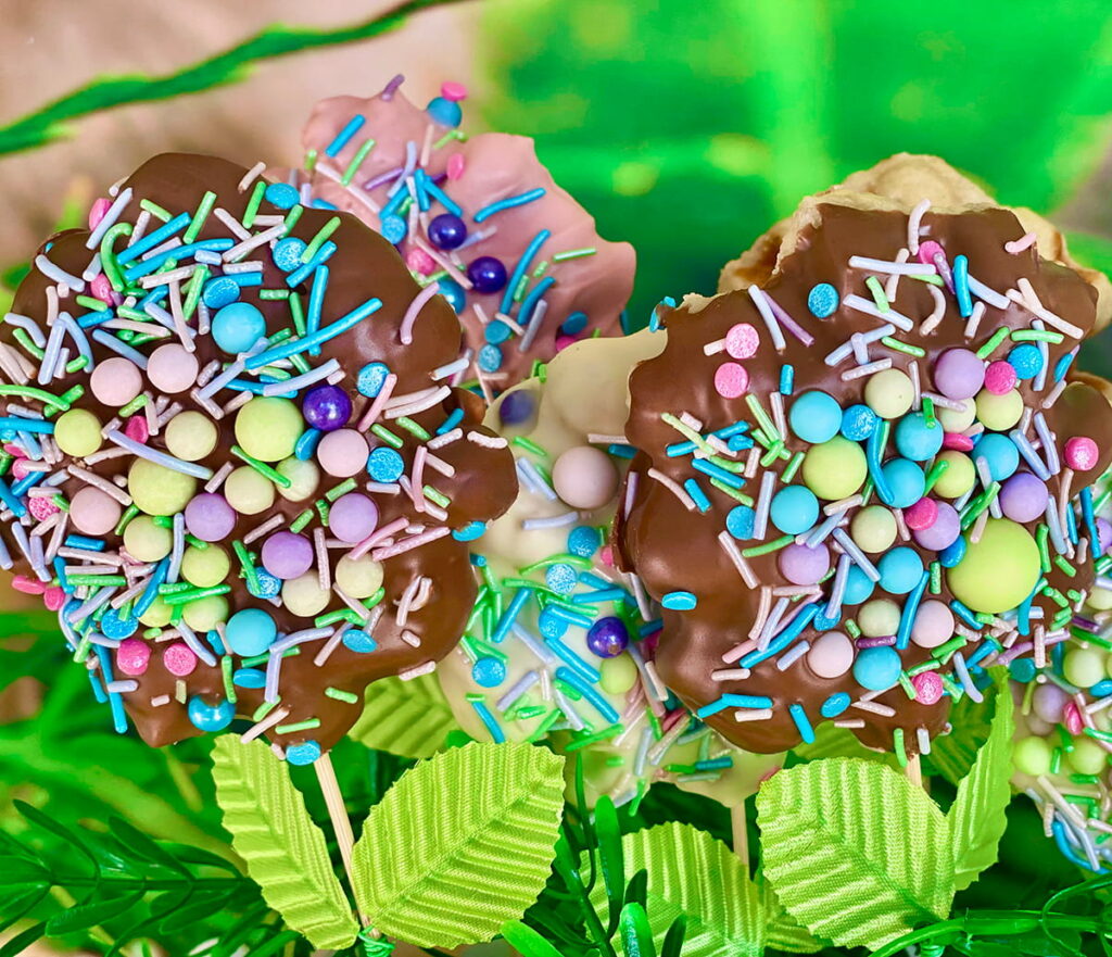 Close-up of five Easter waffle pops on sticks with faux green leaves—front pops dipped in vibrant chocolate and covered in bright sprinkles, with white and strawberry chocolate-covered waffle pops arranged behind.