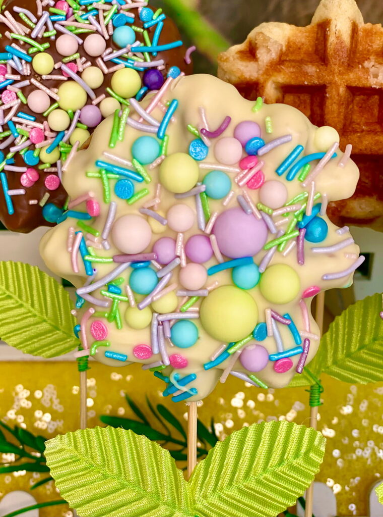 Close-up of three Easter waffle pops on sticks with faux leaves—the front pop is dipped in white chocolate and topped with pastel sprinkles, the second is milk chocolate with sprinkles, and the third is plain.