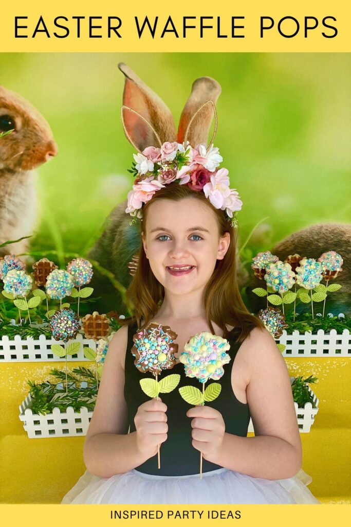 Young girl smiling at the camera while holding two Easter Waffle Pops in front of a spring-themed treat table with a bunny backdrop, yellow sequin tablecloth, and picket-style garden crates displaying waffle pops like spring flowers.