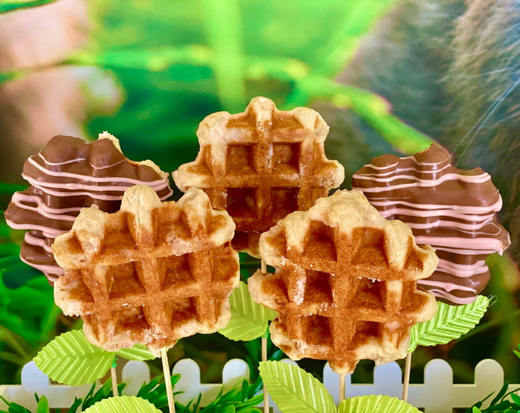 A mix of Easter waffle pops on sticks displayed with faux greenery—three plain and two chocolate-covered pops drizzled with pink chocolate, arranged like flowers for a kids' spring party.