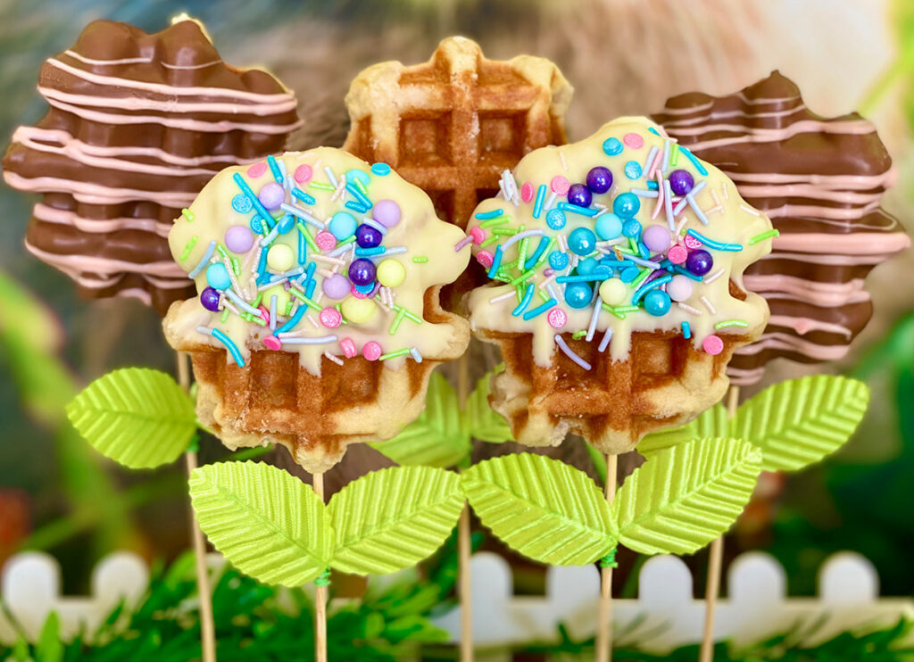 Five Easter waffle pops on sticks styled with faux leaves to resemble blooming spring flowers—two covered in chocolate with pink drizzle, two half-dipped in white chocolate with pastel sprinkles, and one plain.