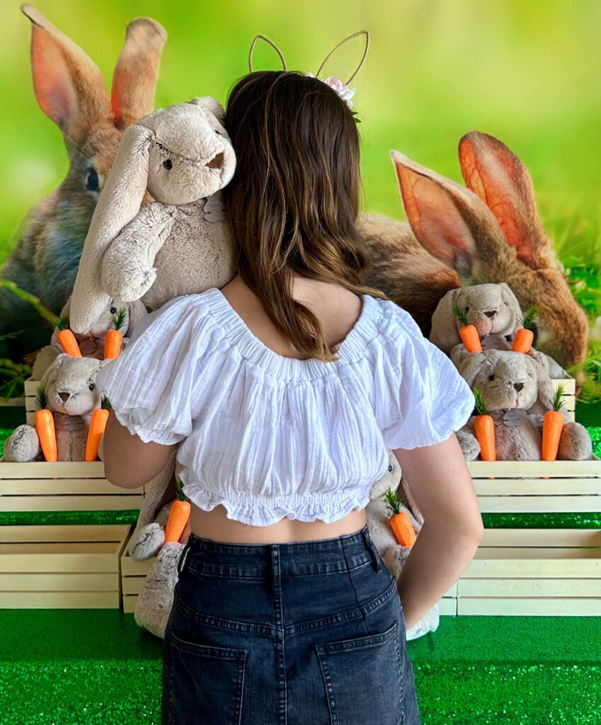 Girl with her back to the camera holds a plush bunny on her shoulder, its face turned toward the lens, in front of an Easter Bunny Adoption table.