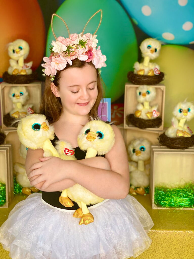 Young girl smiling gently as she looks down at two plush Easter chicks cradled in her arms. A heartwarming and tender moment captured in front of the Easter Chick Adoption party setup.