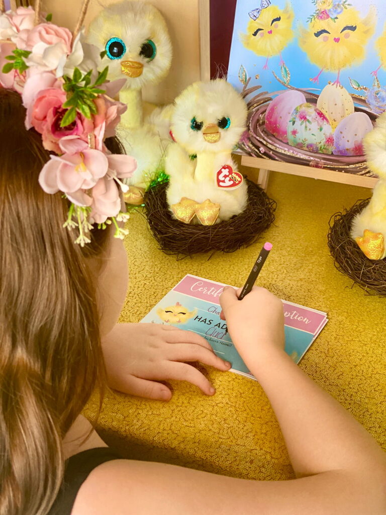 Top-down view of a young girl filling out her Easter Chick Adoption certificate. The back of her head is visible as she writes, with plush chicks and the certificate on the table in front of her.