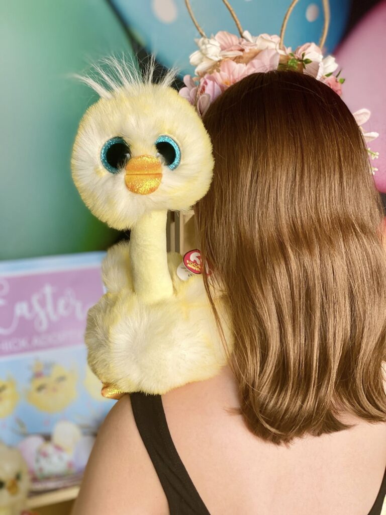Young girl seen from behind, holding a plush Beanie Boo chick on her shoulder, with the chick facing the camera.