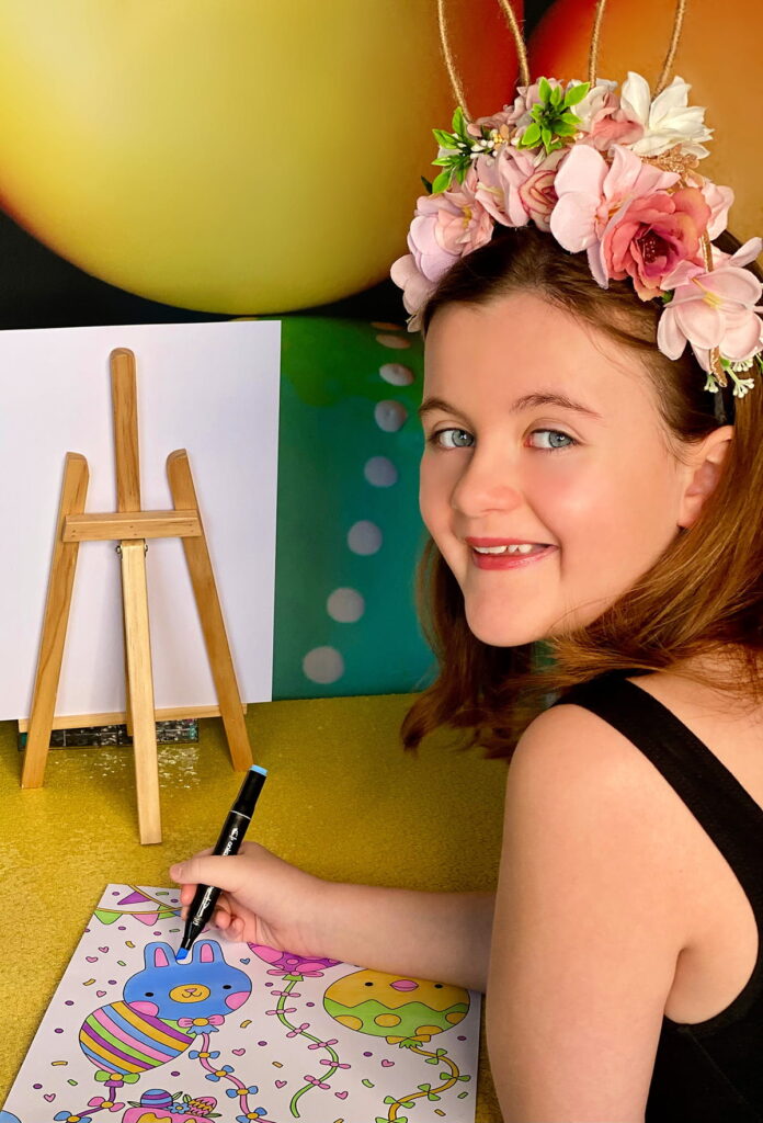 Young girl in floral bunny ears smiling up while colouring at Easter activity table with egg backdrop and yellow sequin tablecloth.