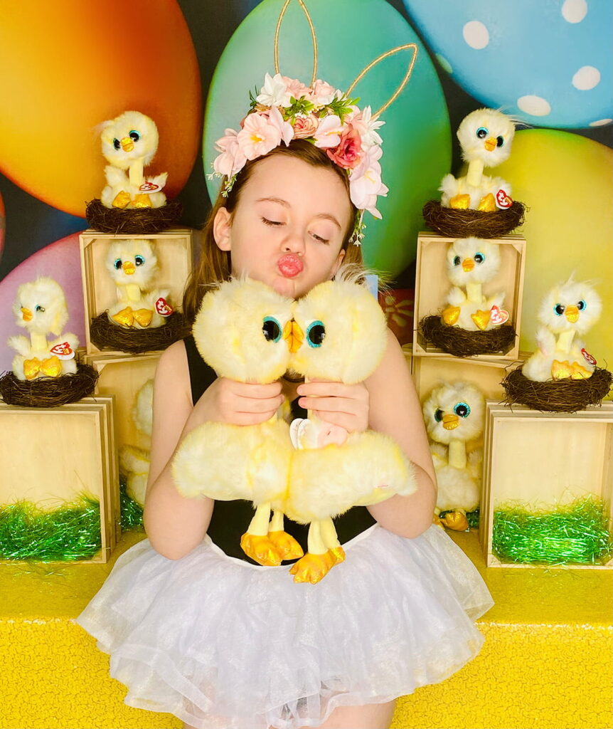 Playful young girl standing in front of the Easter Chick Adoption activity table, holding two plush chicks and making them kiss while making a silly, childlike face. A sweet and innocent moment captured in front of the festive party setup.