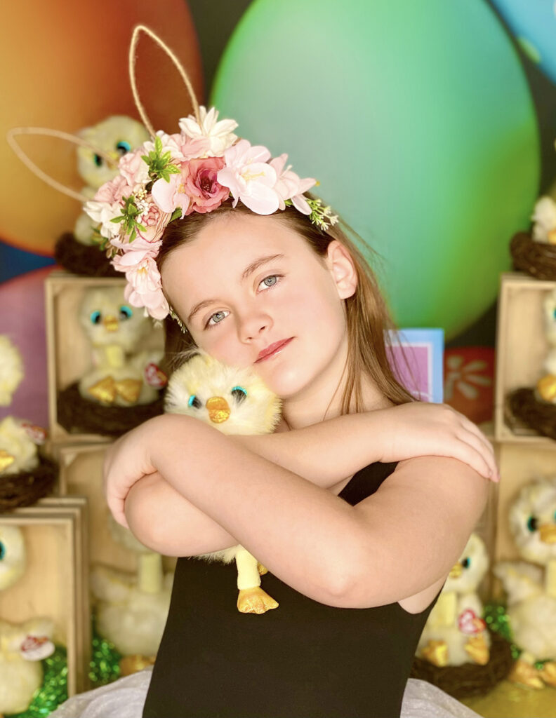 A young girl snuggles a plush Easter chick close to her chest, looking quietly content in front of a colorful adoption activity table.