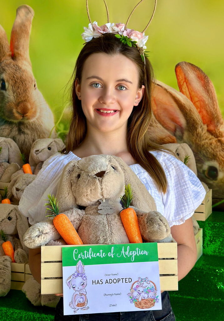 Girl smiling in floral bunny ears headband, holding a wooden crate filled with a plush bunny, carrots, and an Easter Bunny Adoption certificate.
