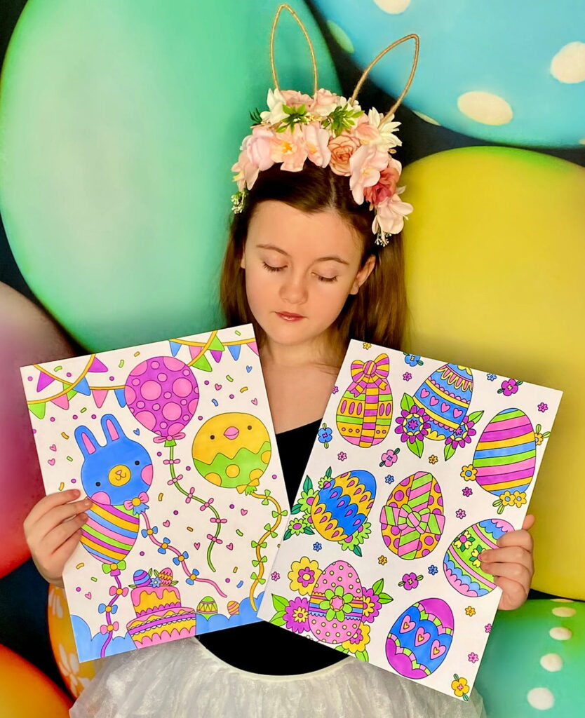 Young girl standing in front of a vibrant Easter egg backdrop, looking down at two Easter colouring posters she has completed as part of the party activity.
