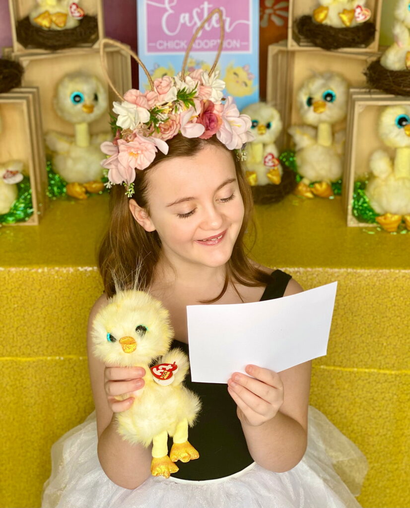 Young girl sitting at the Easter Chick Adoption table, smiling as she holds a plush chick and looks down at her filled-out adoption certificate.