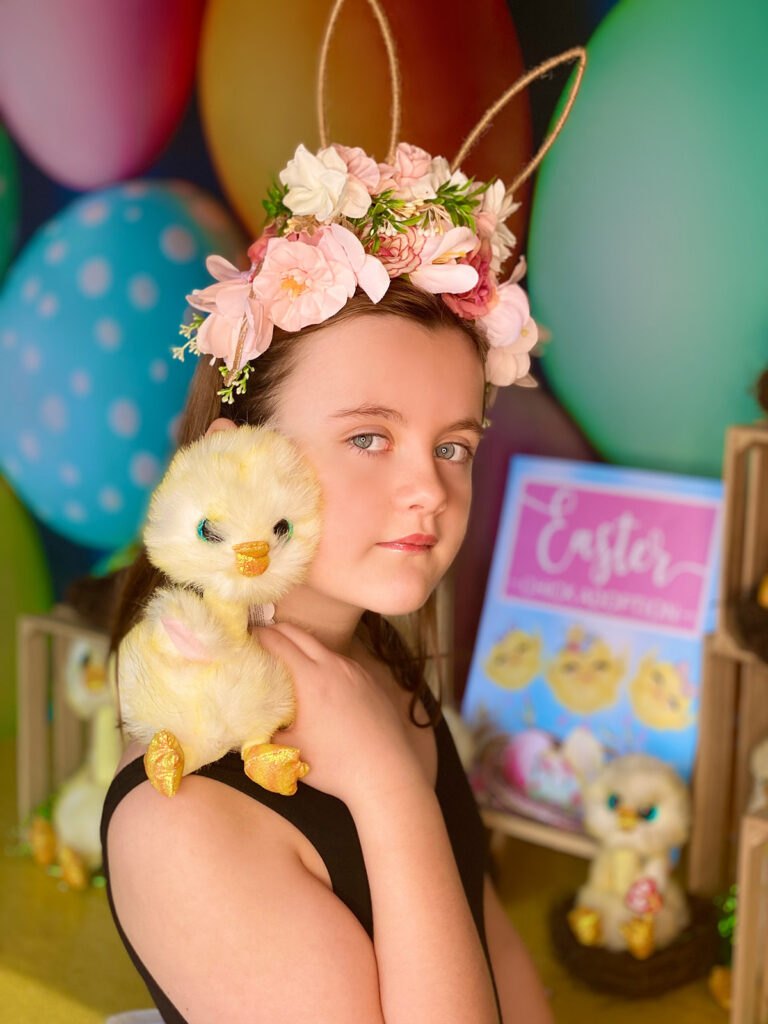 Young girl wearing a floral Easter headband holding a plush Easter chick on her shoulder. Both the girl and chick are looking at the camera, standing in front of the Easter Chick Adoption party table.