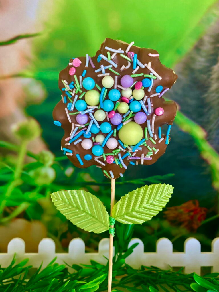 Single Easter waffle pop covered in milk chocolate and pastel sprinkles, styled with faux green leaves and displayed in a white crate with green foliage at the base and a soft spring backdrop behind.