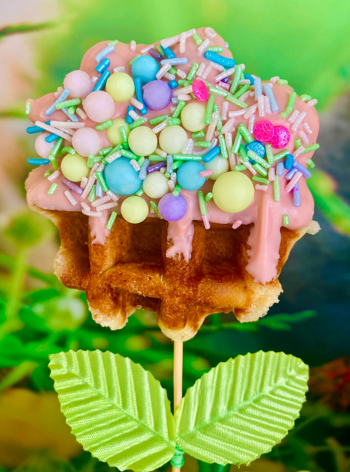 Single Easter waffle pop on a stick with faux leaves, half dipped in pink chocolate and topped with pastel Easter sprinkles, styled to look like a blooming flower.