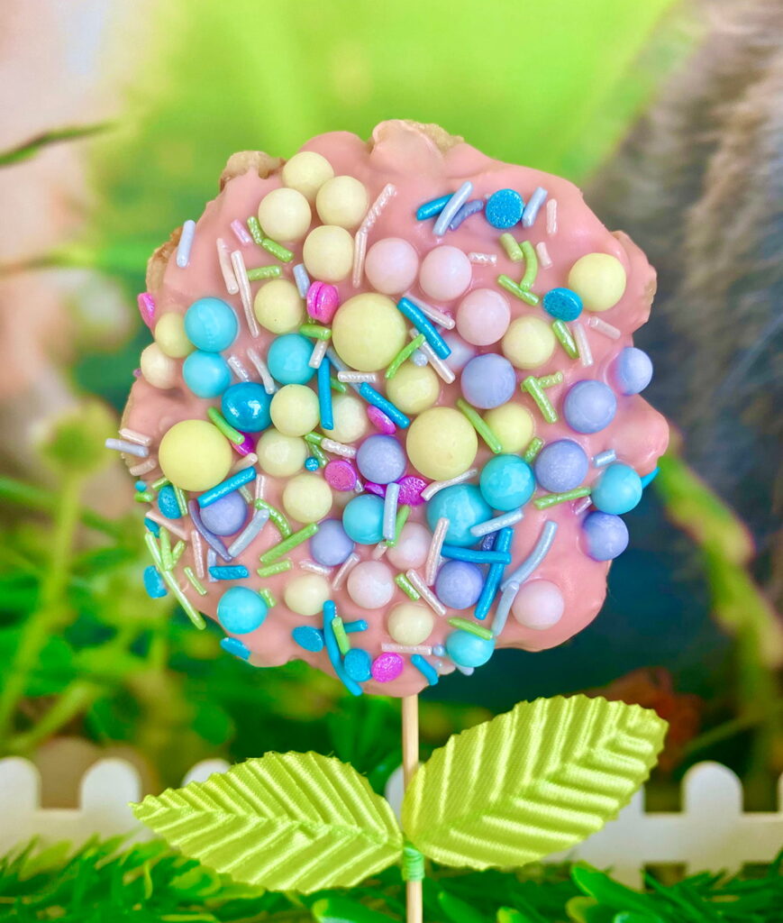 Easter waffle pop fully covered in pink chocolate and topped with pastel sprinkles, displayed with faux green leaves to resemble a festive spring flower.
