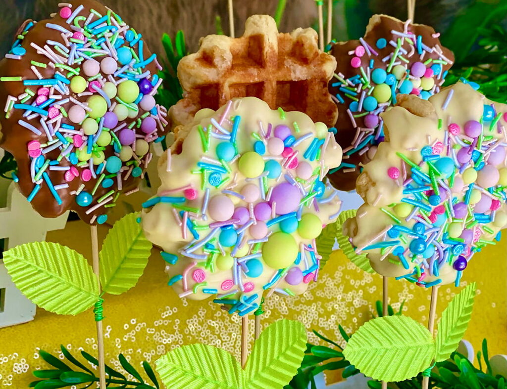 Close-up side angle of a kids’ Easter waffle pops table featuring chocolate-dipped and white-dipped waffle pops covered in pastel sprinkles, alongside one plain waffle pop.
