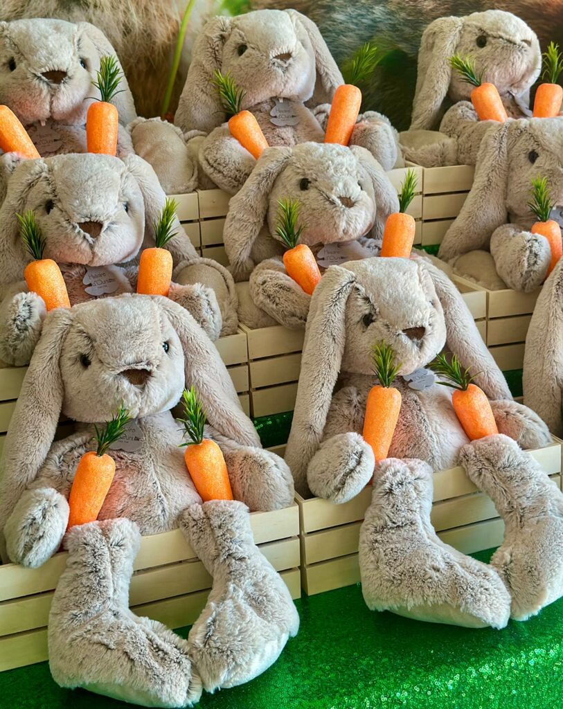 Side angled view of plush bunnies sitting in wooden crates with faux carrots as part of an Easter-themed display for a kids party.