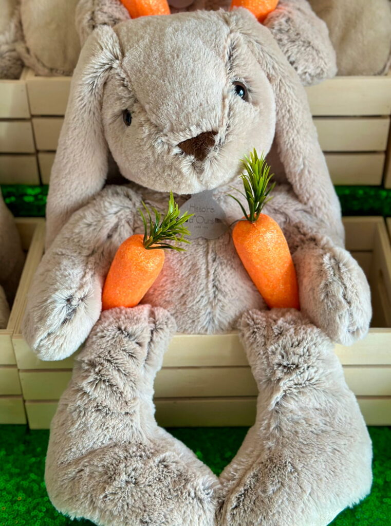 Front top-down view of a plush bunny sitting in a wooden crate with its legs hanging over the edge and a faux carrot placed on each side.