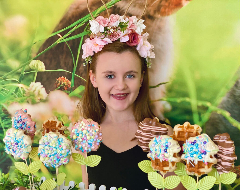 Smiling young girl wearing a floral bunny ear headband standing behind a colorful Easter waffle pops table with a bunny-themed backdrop.