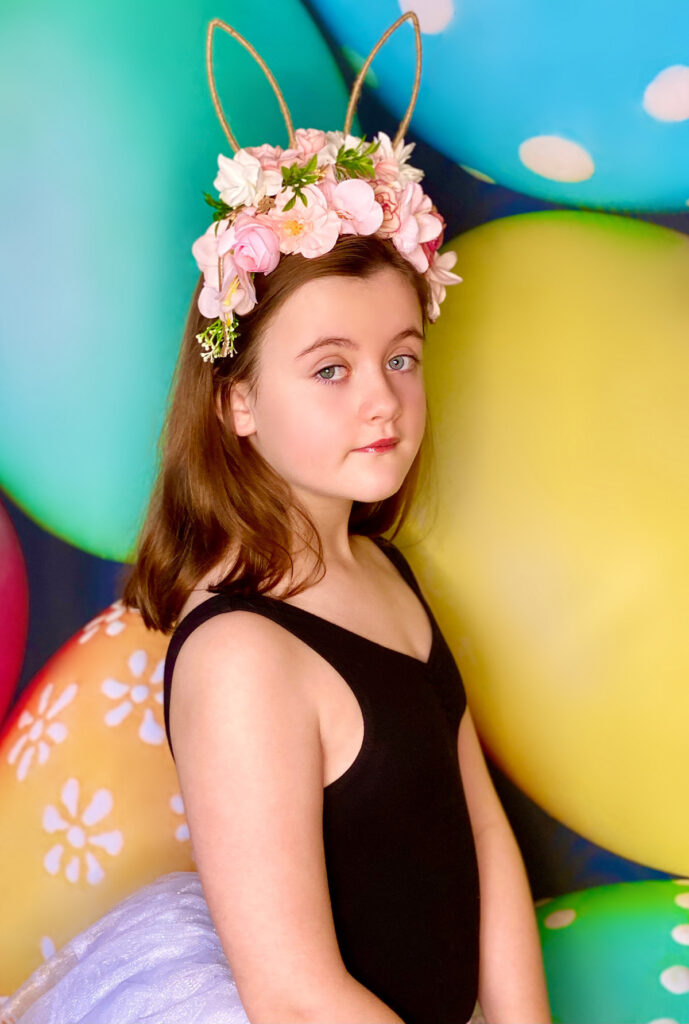 Young girl wearing a floral bunny ear headband standing in front of a vibrant egg-themed Easter backdrop, looking at the camera with a sweet, innocent expression.