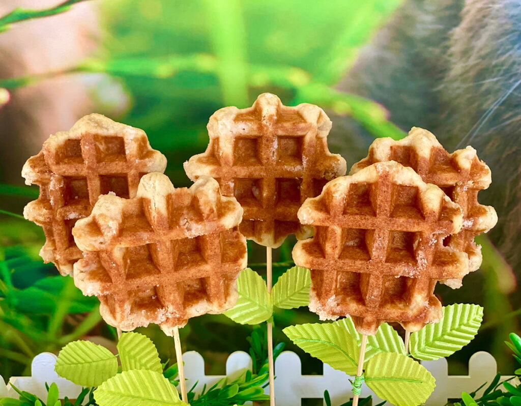 Undecorated Easter waffle pops on sticks arranged with faux leaves to look like blooming flowers, styled as a spring-themed kids' party treat with a softly blurred background.