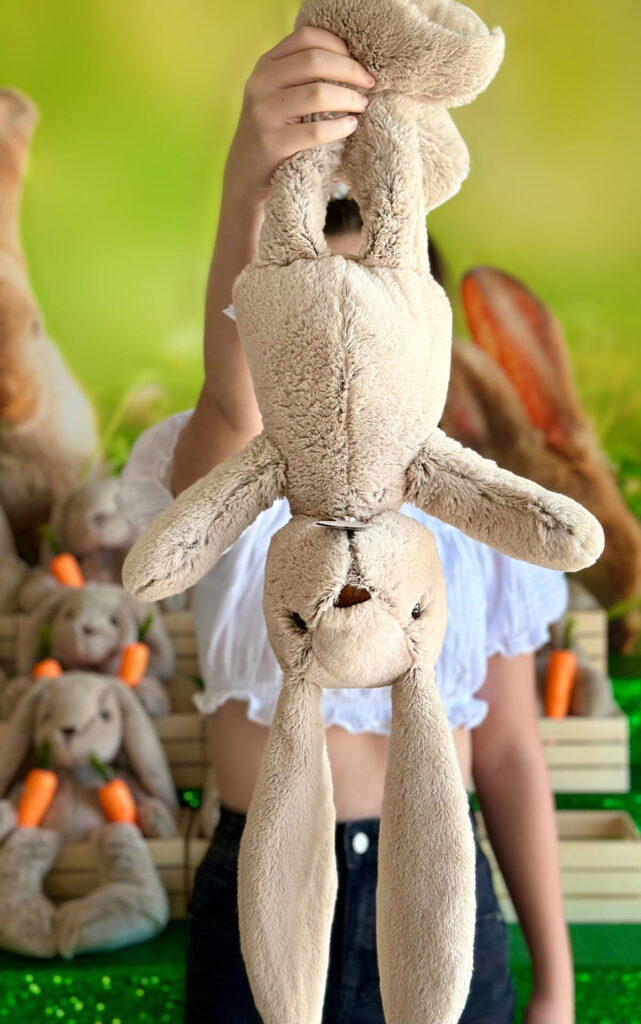 Plush bunny held upside down by its feet, with long ears hanging down in a playful close-up shot; the child holding it is mostly out of frame.