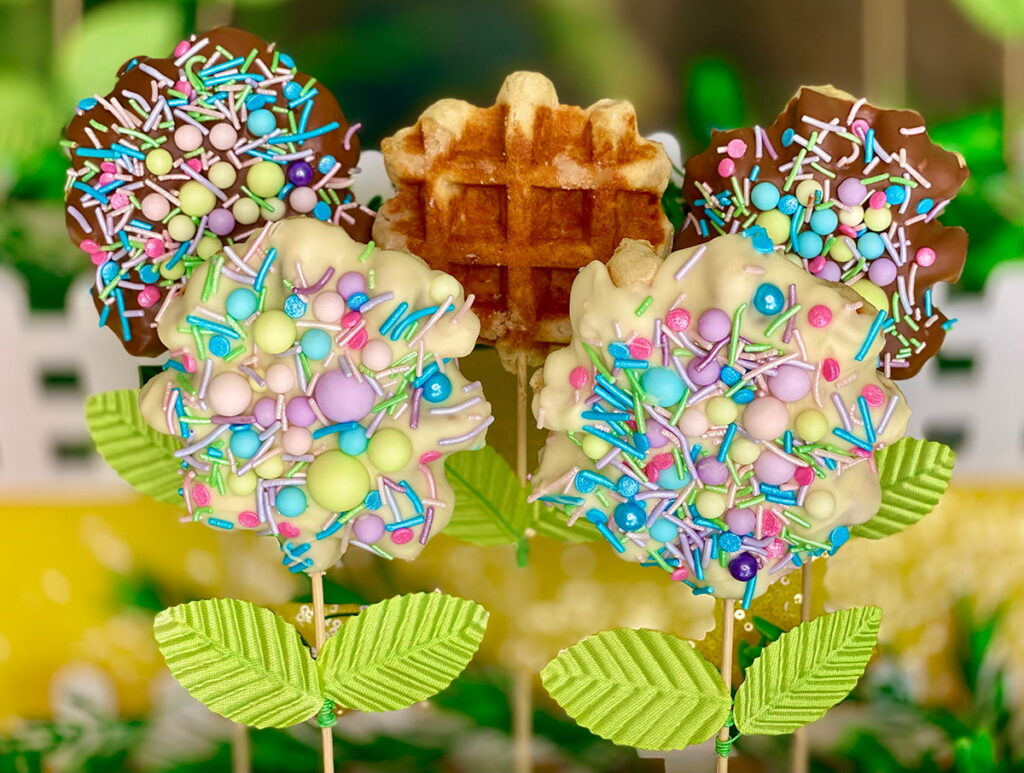 White chocolate and milk chocolate Easter waffle pops covered in pastel sprinkles, styled with faux leaves to look like spring flowers, perfect as a kids’ Easter party treat idea.