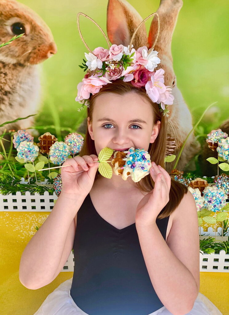 Young girl wearing a floral bunny headband biting into a half white chocolate-dipped Easter waffle pop covered in pastel sprinkles, standing in front of the waffle pops table.