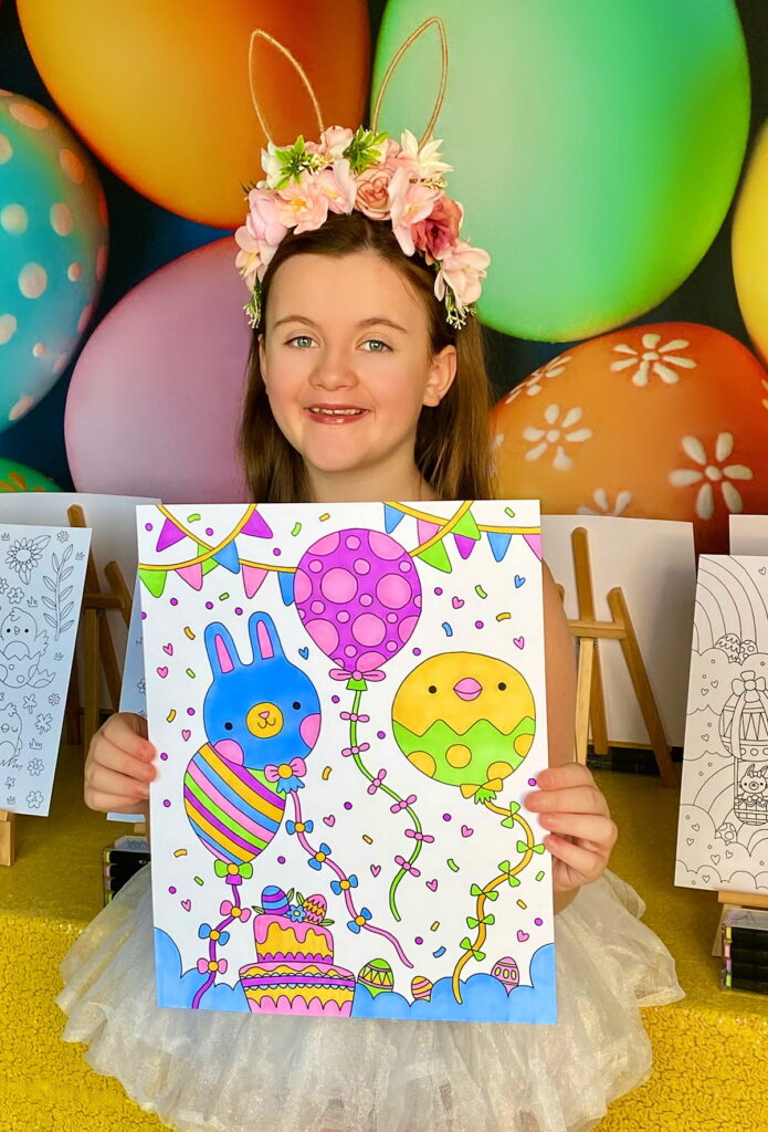 Girl in floral bunny ears and white tutu smiling, holding coloured poster at Easter colouring party with egg backdrop and yellow sequin table.
