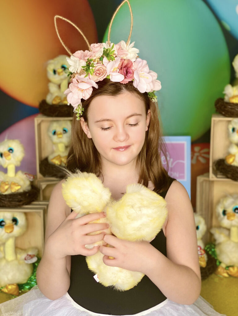 Young girl wearing a floral Easter ear headband, looking down sweetly and innocently at two plush chicks she is holding in her hands, with the Easter chick activity table blurred behind her.