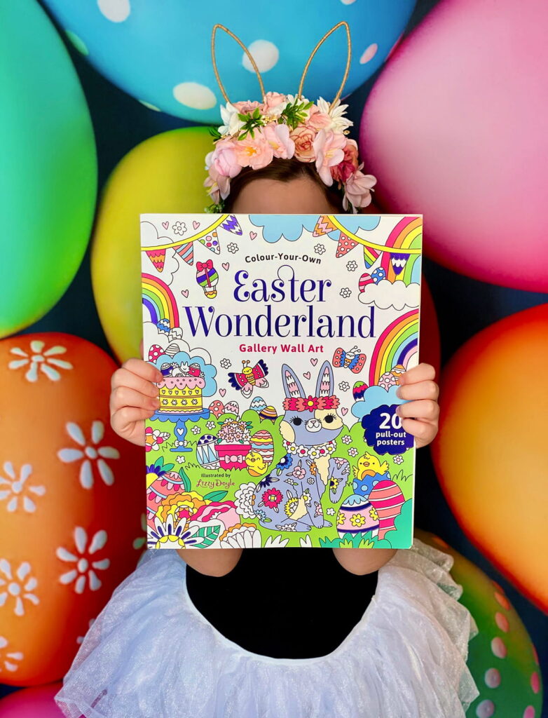 Young girl standing in front of a vibrant Easter egg backdrop, holding an Easter wall art book filled with uncoloured posters for the kids’ colouring activity.