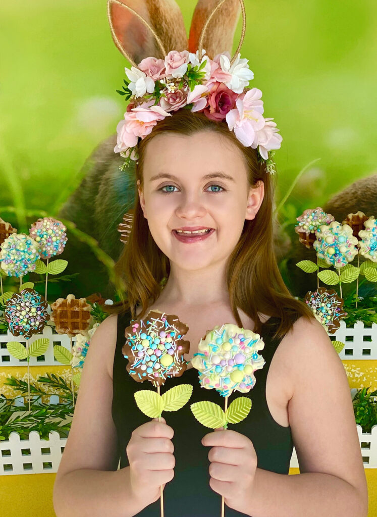 Young girl wearing a white tutu and floral bunny ear headband, standing in front of an Easter waffle pops table while holding two waffle pops—one chocolate and one milk chocolate, both covered in pastel sprinkles.