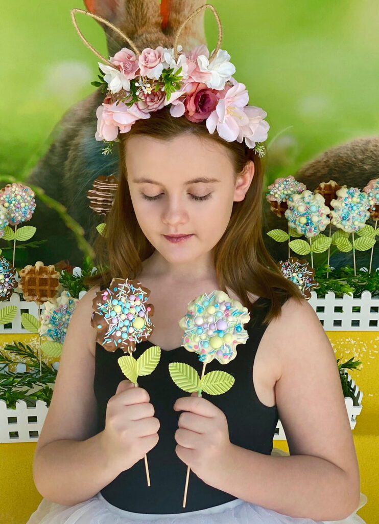Young girl holding two Easter waffle pops—one milk chocolate and one white chocolate—looking down at them while standing in front of the waffle pops table.