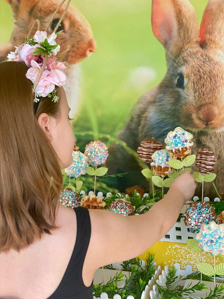 Back of young girl’s head wearing a floral headband as she reaches to pick up a waffle pop from an Easter waffle pop display, featuring a spring bunny backdrop, yellow sequin tablecloth, and white picket-style crates.