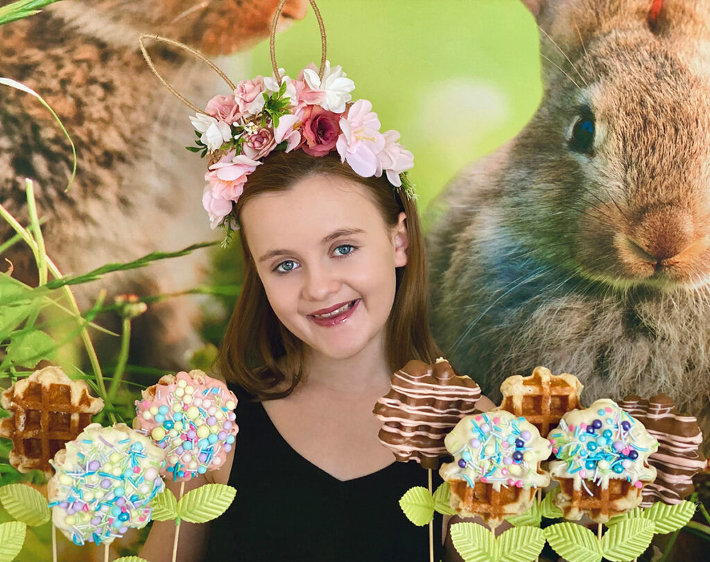Young girl wearing a floral bunny ear headband, smiling behind a table of decorated Easter waffle pops with a festive bunny backdrop in the background.