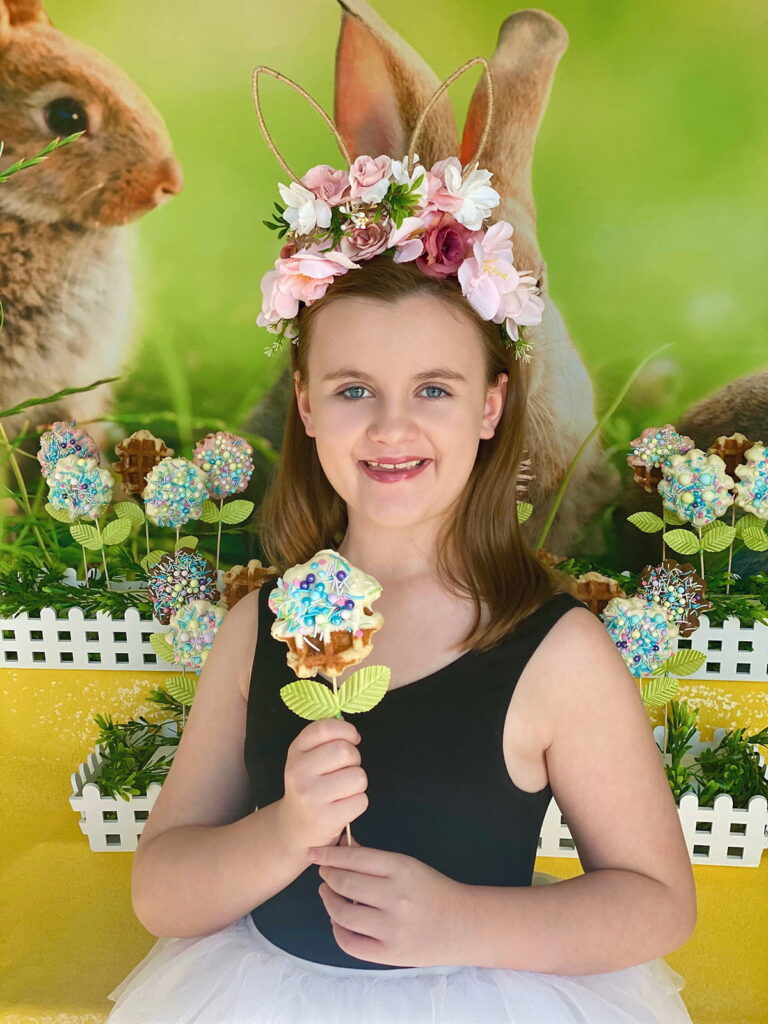 Young girl smiling happily while holding a white chocolate-dipped Easter waffle pop covered in pastel sprinkles.