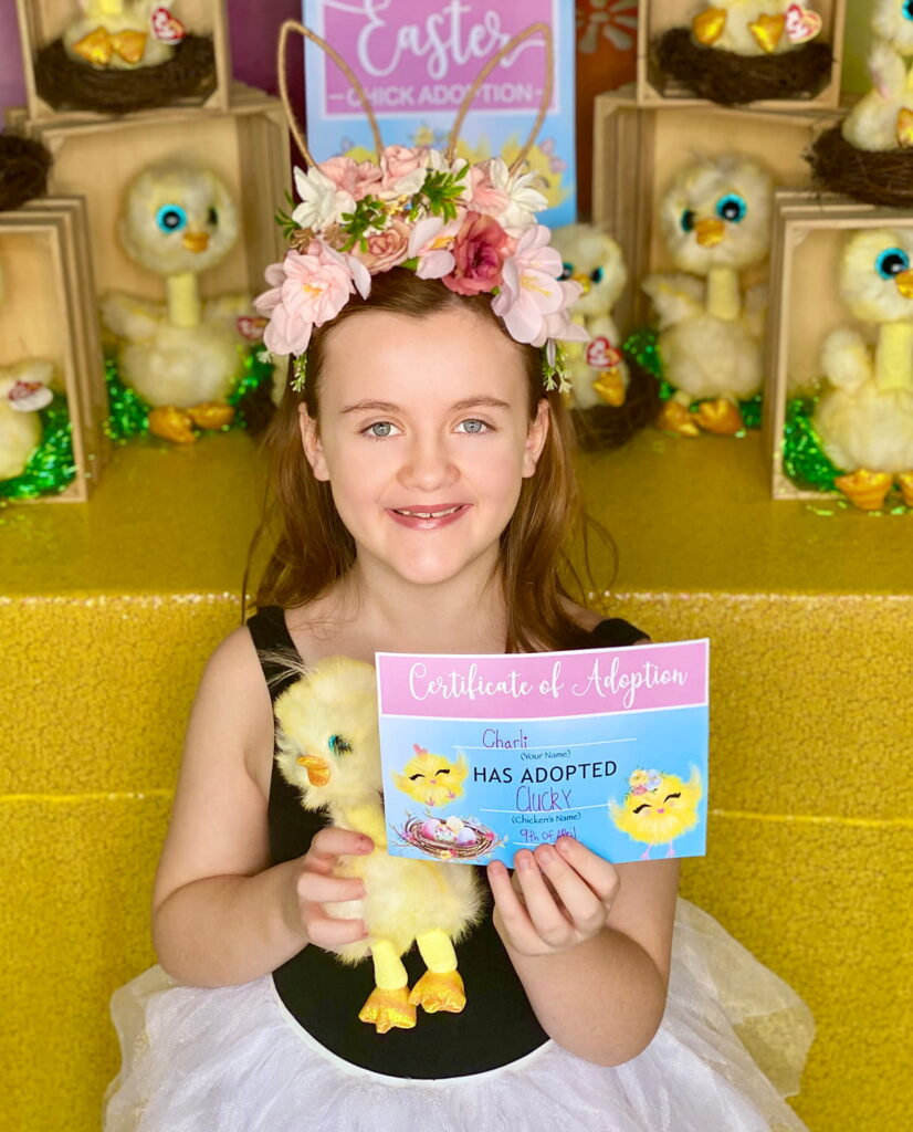Young girl smiling while seated in front of the Easter Chick Adoption activity table, holding her plush Beanie Boo chick and a completed adoption certificate.
