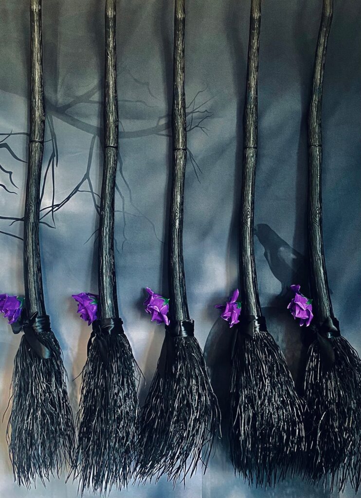Five black witch brooms topped with purple roses, displayed before a spooky backdrop for a DIY witch broom decorating party.