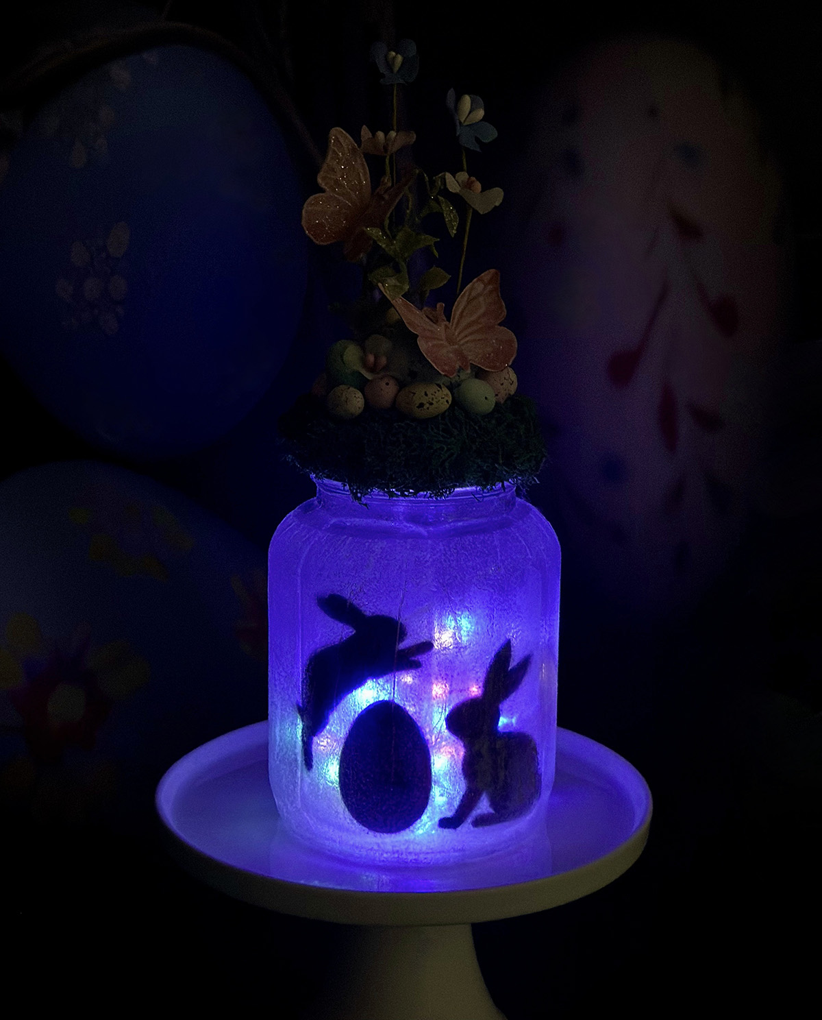 Decorated Easter Fairy Jar Lantern on a cake stand glowing with blue-hued fairy lights, featuring a bunny jumping over an egg to another sitting bunny, with butterflies and a floral moss lid topper visible, set in a darkened scene to highlight the illuminated jar.