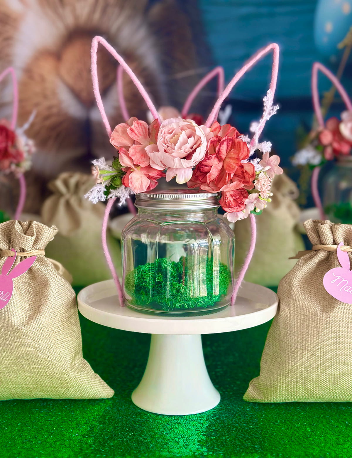 Close-up of a clear mason jar on a white cake stand with a floral bunny ear headband decorating the lid, bright green moss inside the jar, and hessian bags filled with supplies showcase the individual setup for each child at the Easter terrarium party.