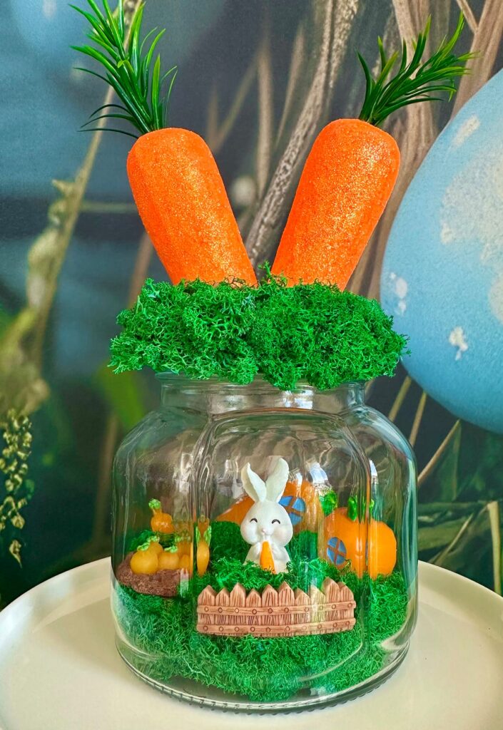 Close-up of a decorated Easter terrarium on a white cake stand, featuring a moss-covered lid topped with two foam carrots and a charming Easter scene inside with miniature carrot and bunny decorations.