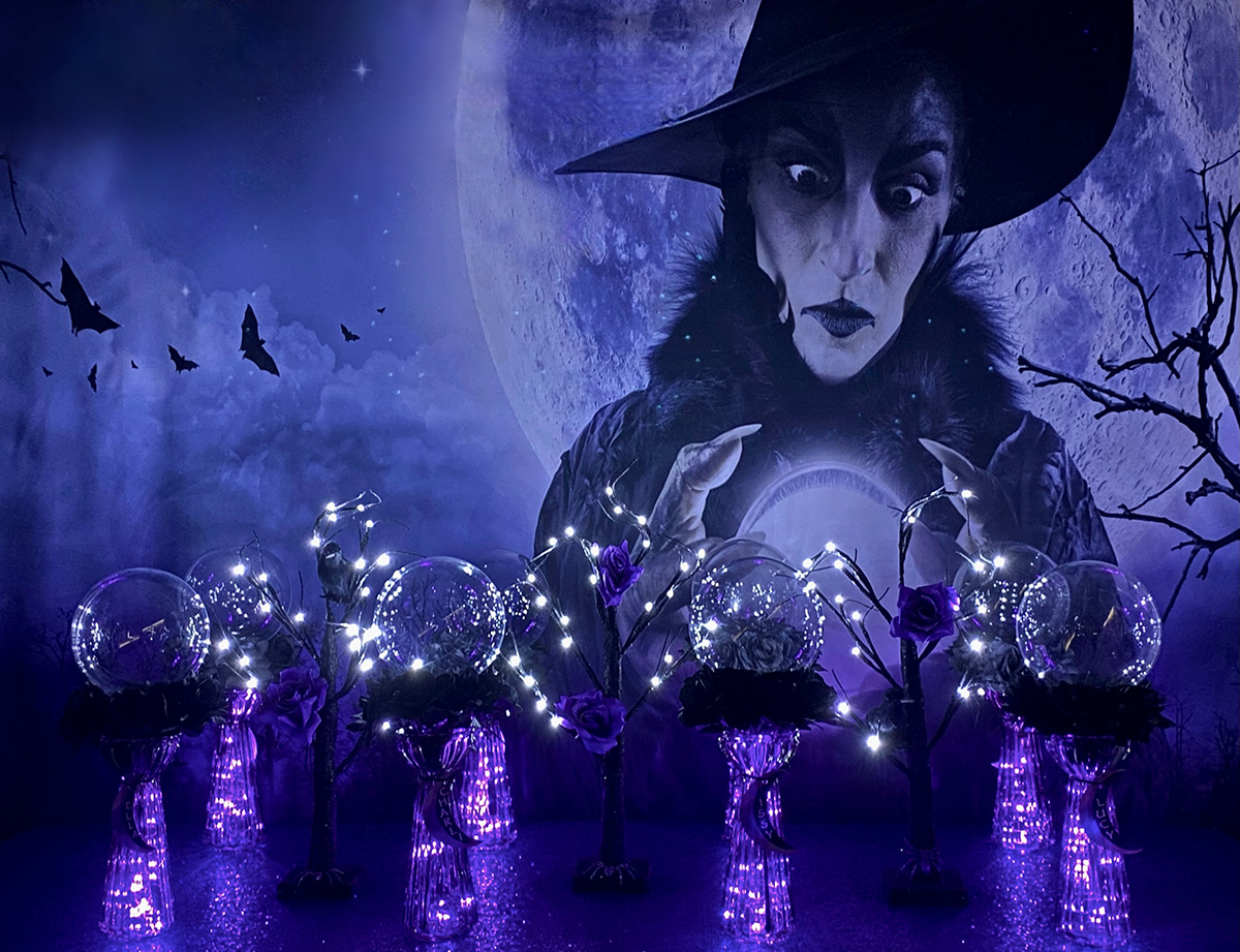 DIY crystal ball party table setup with acrylic spheres resting on glass vases filled with purple fairy lights, surrounded by LED trees decorated with flowers and crows, in front of a spooky witch backdrop with bats and a full moon.