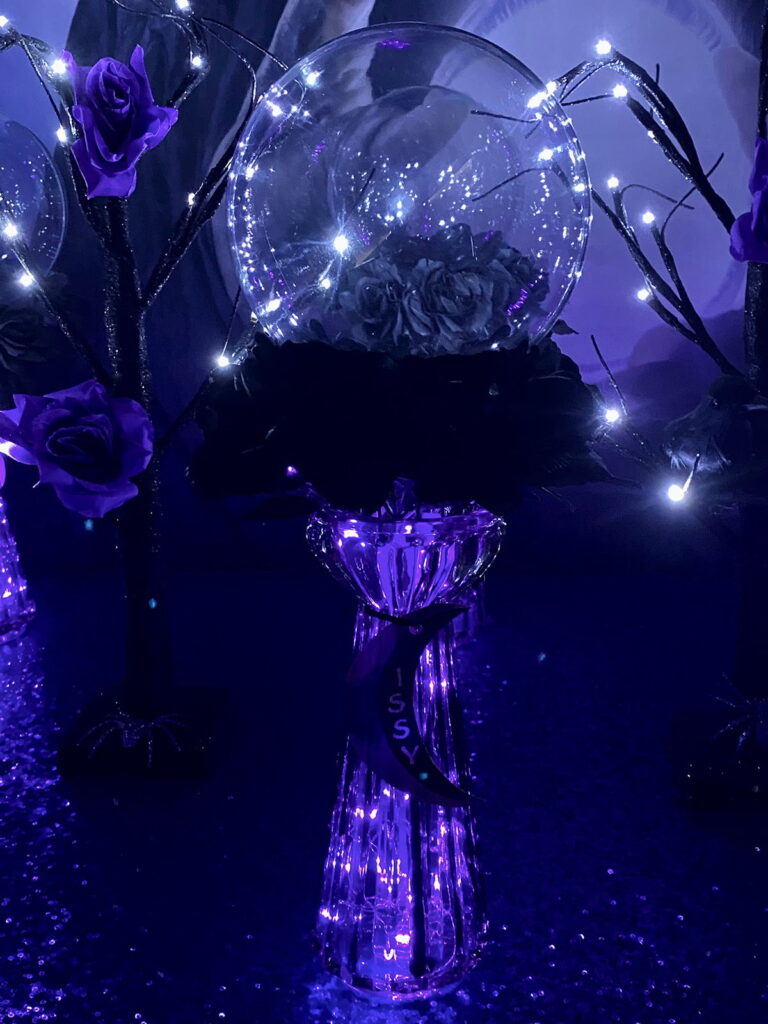 Ready-to-decorate acrylic crystal ball on a glass stand with glowing purple fairy lights and black flowers, for a kids’ witch party activity.