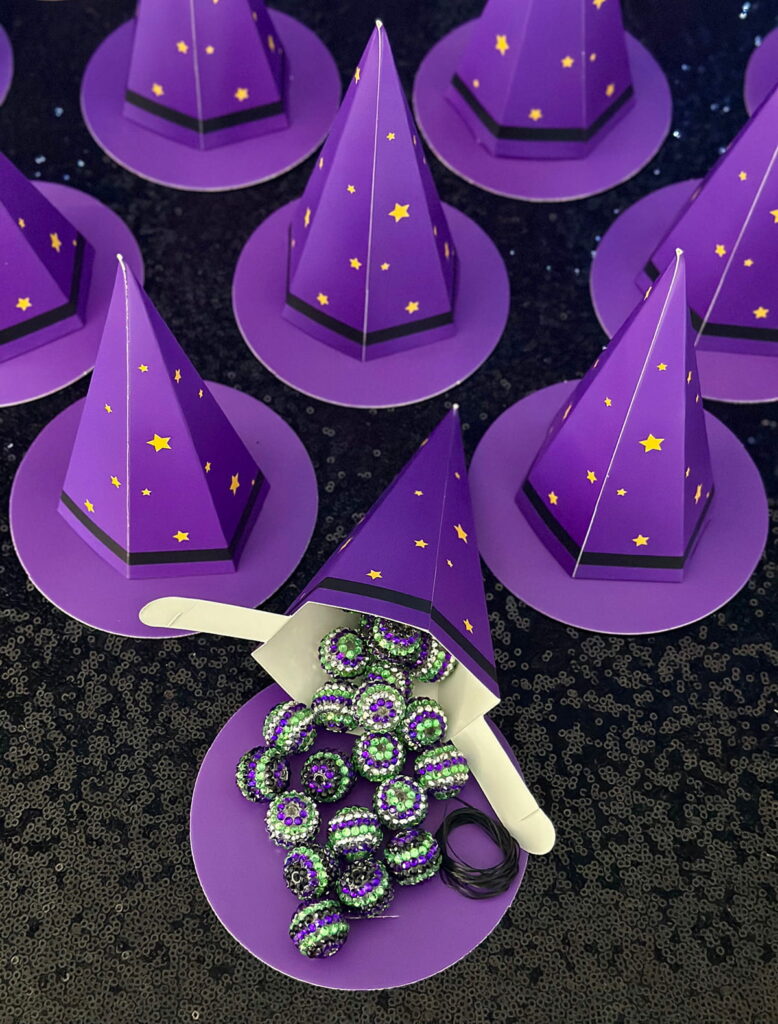 Witch-themed party favour with cardboard purple witch hats decorated with gold stars; one hat is open, spilling purple, black, and green themed beads to reveal a DIY jewelry kit inside, perfect for a kids’ witch-themed party.