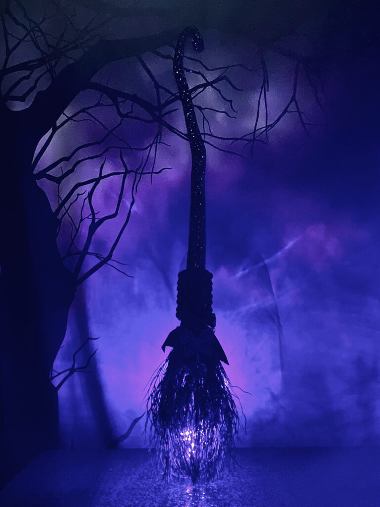 Black-decorated DIY witch broom lit with purple LED lights, displayed in front of a spooky backdrop, creating an eerie and magical glow.