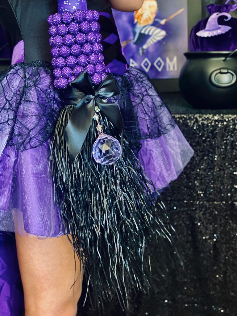 Close-up of a DIY witch broom decorated in black and purple with a hanging crystal, part of a kids’ witch-themed party activity.