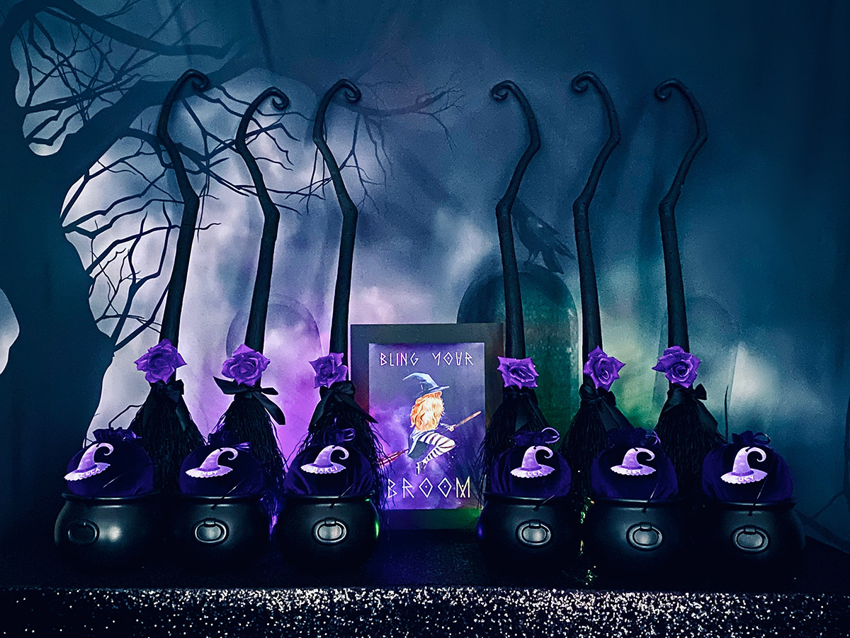 DIY witch broom decorating party setup with black brooms topped with purple roses in cauldrons, on a black sequin tablecloth before a spooky graveyard backdrop.