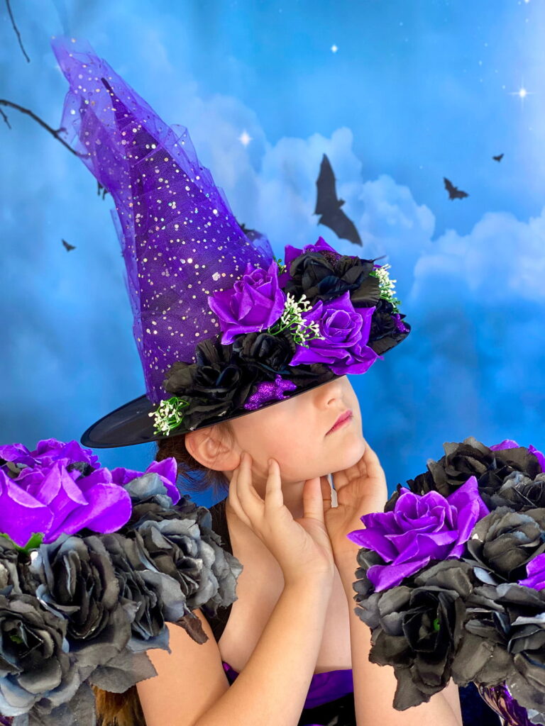 Young girl wearing a DIY purple-themed witch hat decorated with purple and black flowers and tulle, perfect for a kids’ witch-themed party.